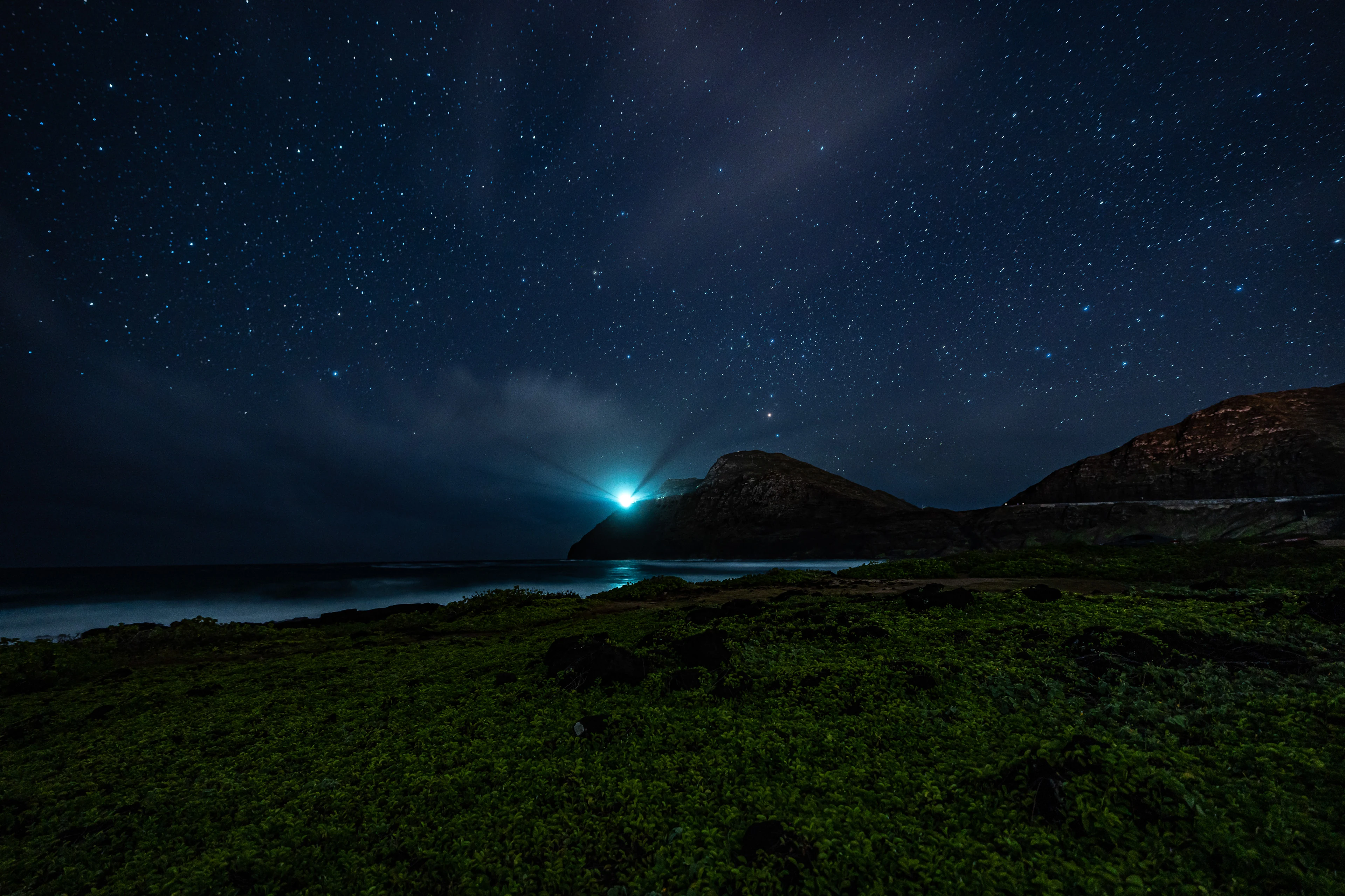 A blue light beyond a coastal ridge beneath a starry sky