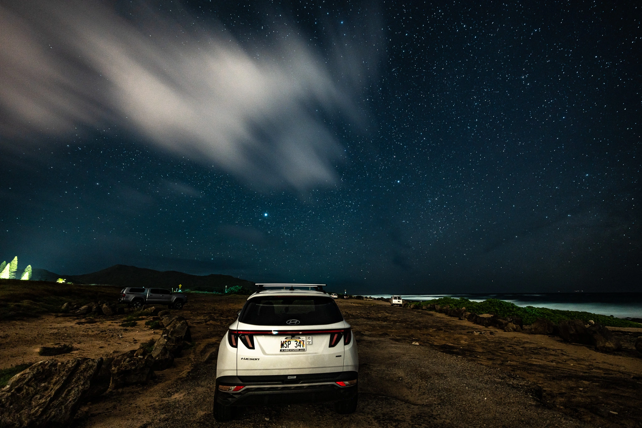 A white car parked beside the ocean beneath stars and fast-moving clouds