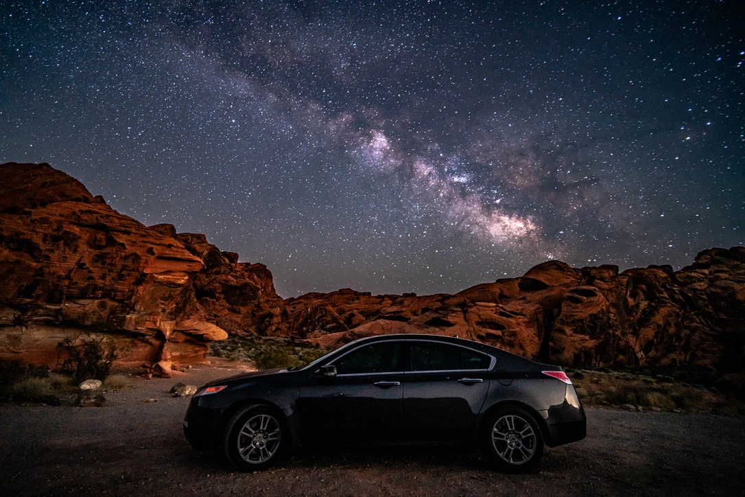 A black car beneath the Milky Way among red desert rocks